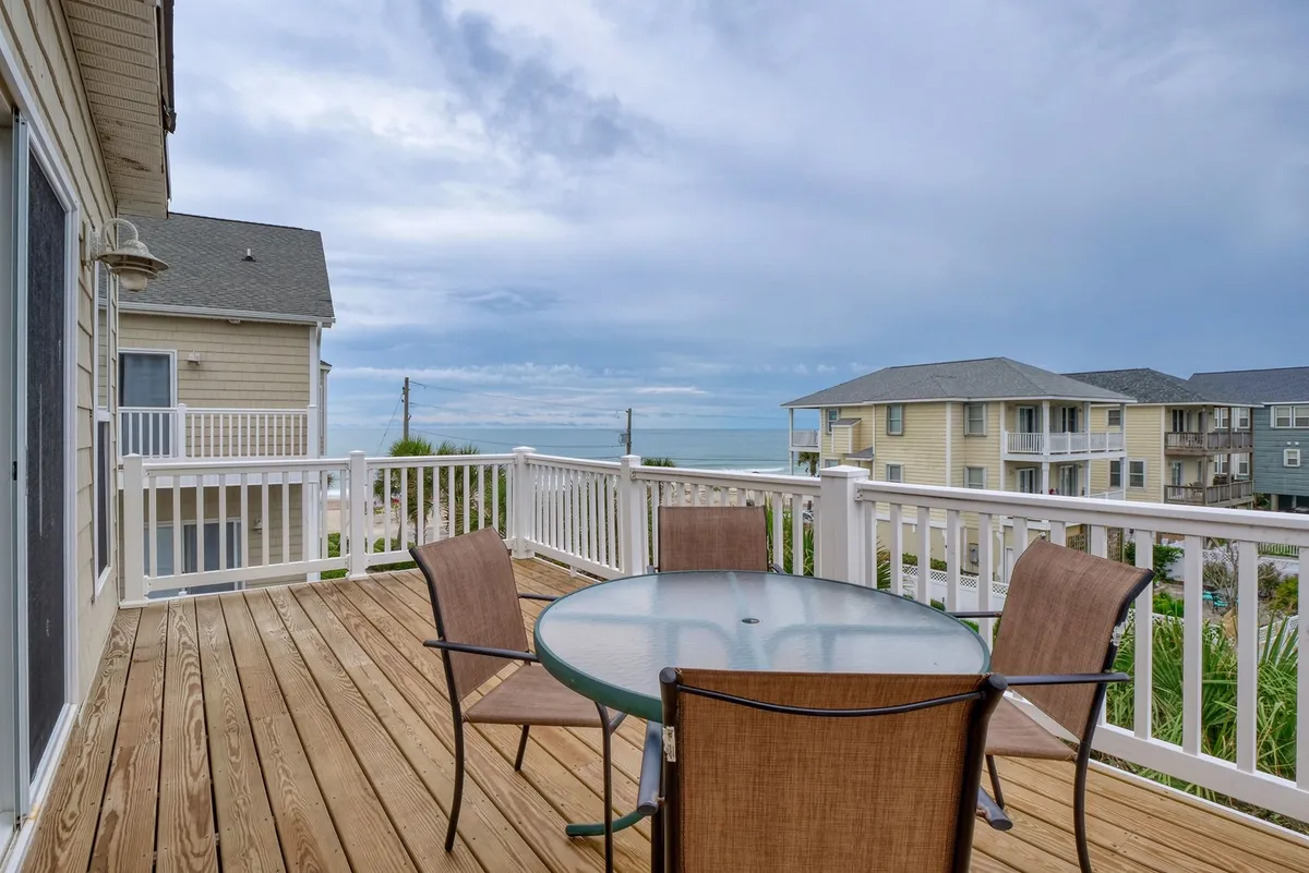 The image showcases a spacious wooden deck overlooking the ocean, featuring a round glass table with four inviting brown chairs—perfect for outdoor dining or relaxing. The scene exudes a casual, coastal charm, with clear views of the water and sky, hinting at the tranquil experience guests can enjoy from sunrise to sunset.