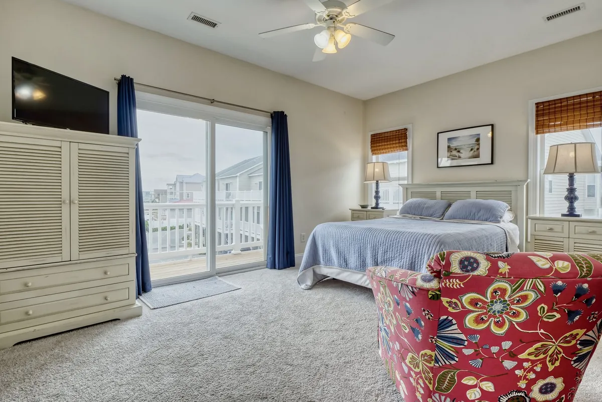 This interior image shows a comfortable bed with a colorful chair nearby, illuminated by natural light pouring through sliding doors that open onto a balcony. The setup invites guests to relax, read, or step outside to enjoy the view.