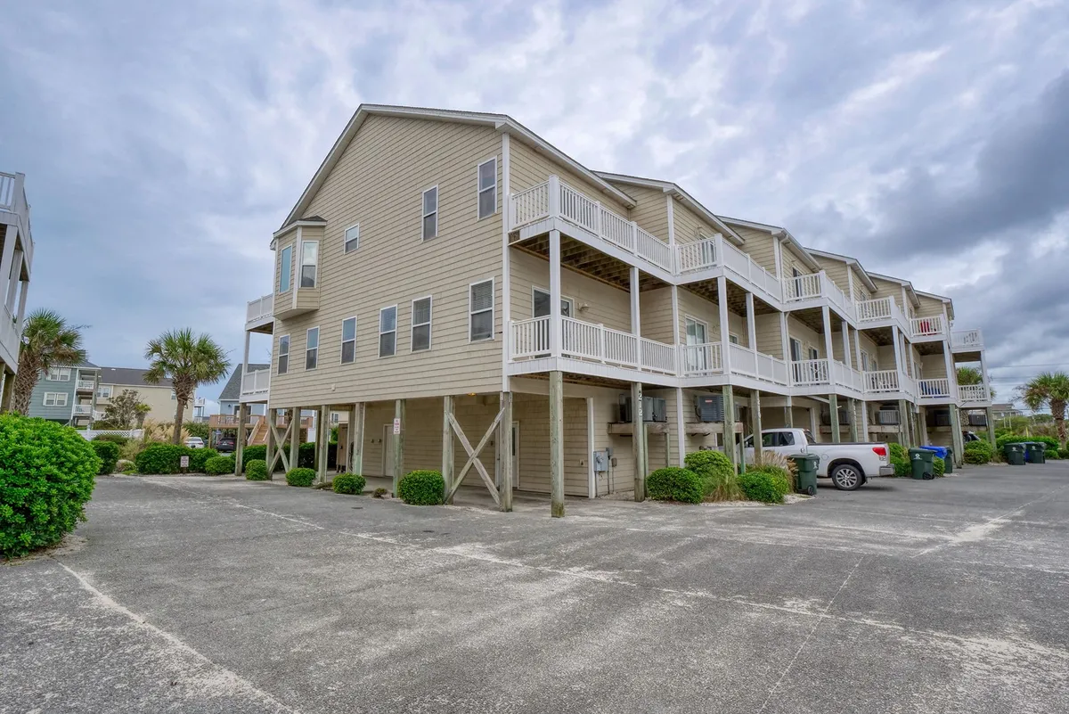 A two-story building with balconies is surrounded by swaying palm trees and a neatly maintained parking area. The balconies offer a view over the local area, suggesting easy access to neighborhood shops, eateries, and the pier. The scene feels inviting, with hints of sunset light and casual outdoor activity.