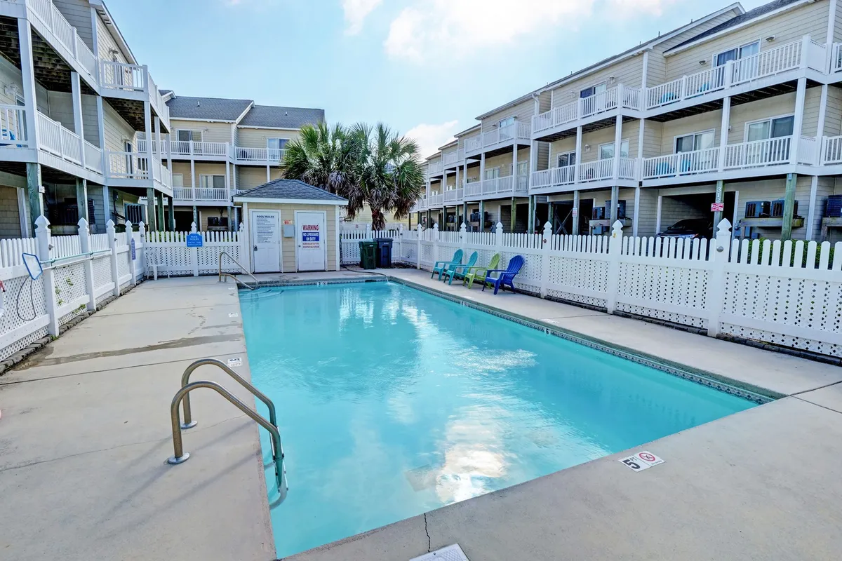 A bright, inviting swimming pool is the centerpiece of this outdoor space, surrounded by secure fencing and bordered by comfortable lounge chairs. Multiple balconies overlook the pool, hinting at easy access from nearby accommodations. The scene captures the essence of community gathering, with spaces for swimming, sunbathing, and socializing.