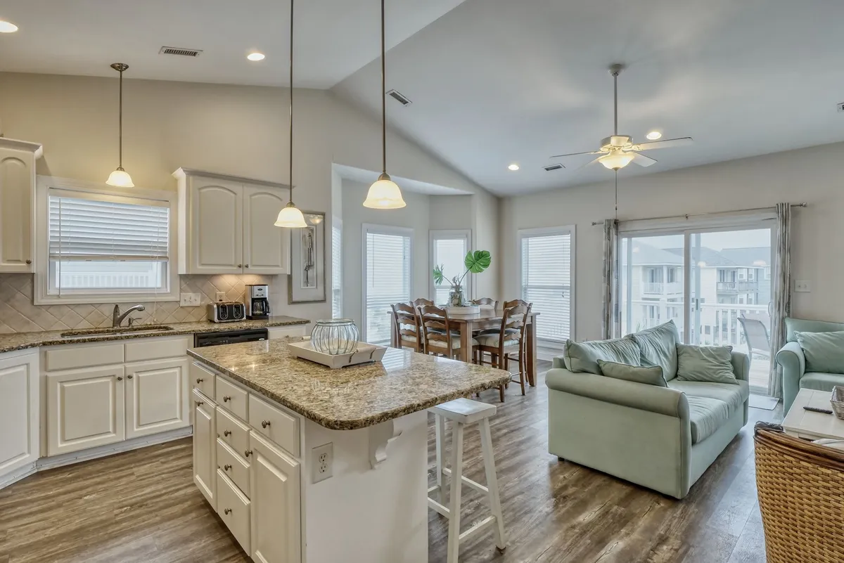 This image presents a wide view of the open-plan layout, featuring the kitchen island with bar seating, a dining area, and comfortable living room seating in one cohesive, sunlit space. The kitchen showcases granite countertops and stainless steel appliances, while the dining and living spaces are arranged for easy interaction and group gatherings.