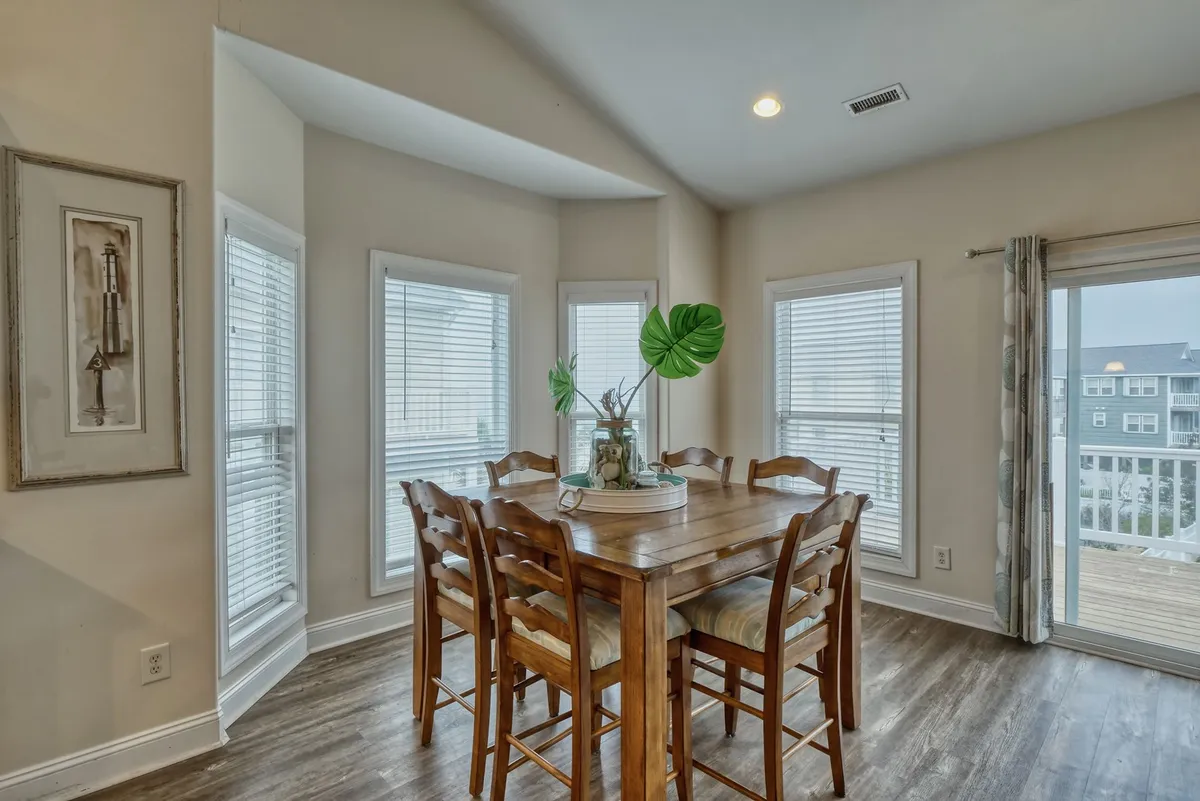 A bright dining area is showcased with a large table, surrounded by windows that let in ample natural light and offer glimpses of the outdoors. The space is decorated simply, with a plant adding a touch of freshness, and suggests opportunities for shared meals with a view.