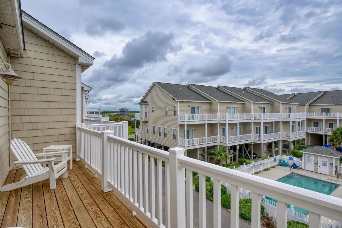 A peaceful balcony overlooks the pool area, furnished with white chairs and surrounded by the neighboring buildings. The pool below is visible, providing a sense of proximity and accessibility from your accommodation. The view evokes a sense of relaxation and connection to the resort environment.