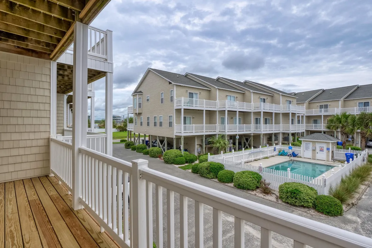 This exterior image showcases a balcony overlooking a sparkling community swimming pool surrounded by landscaped greenery and neighboring buildings. The inviting water, visible poolside activity, and casual seating capture the sense of a lively, shared outdoor amenity area.